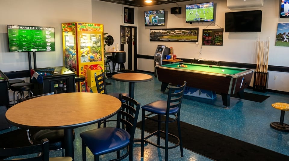 A game room with a pool table, arcade games, high-top tables, and TVs showing sports.