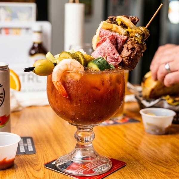 A Bloody Mary cocktail garnished with shrimp, olives, pickles, and a skewer of mini sandwiches.