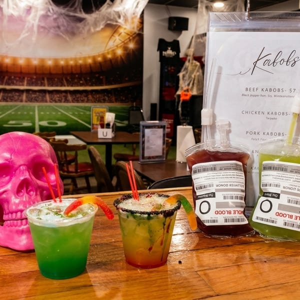 Halloween-themed drinks and decor, including a pink skull, on a wooden bar with a football stadium mural.